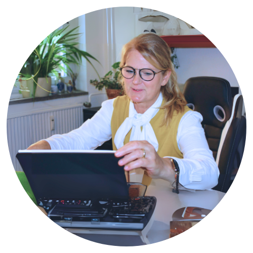 Profilbild von Frau Carmen Blenn-Tietz. Sie trägt Brille, eine weiße Bluse mit einem sonnengelben Cardigan und schaut hinunnter zu ihrem Laptop am Schreibtisch.